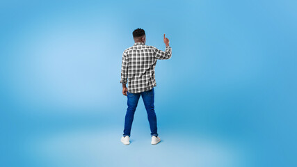 Wall Mural - Back view of young black guy pushing button on virtual screen over blue studio background, mockup for design