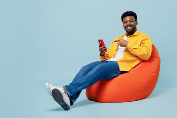 Full size young man of African American ethnicity 20s in yellow shirt sit in bag chair hold in hand use point finger on mobile cell phone isolated on plain pastel light blue background studio portrait