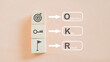 © Ratana21 - OKR dark brown text (Objectives, Key and Results) with dartboard, key and flag icon on wooden cube blocks on pink background  for business planning and measure success, target achievement