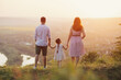 Family - father, mother and little daughter standing on the hill with their backs to the camera and looking to the river and countryside on the sunset.