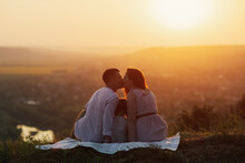 Husband Kisses His Wife While They Sitting On The Hill At Sunset. Little Daughter Sits Between The Parents. Happy Young Family Is Resting In The Countryside. A Bright Sunny Sunset.