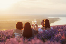 Lovely Ladies Drinking Wine In Lavender Violet Field At Sunset. Summer Happy Mood. Girlfriends Relaxing On Summer Sunset With River On The Background.