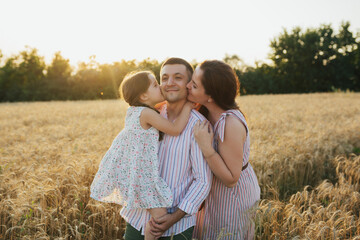 Young mother and her daughter in summer dresses standing  at the wheat field on a sunny day and kissing the happy young father.