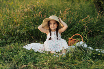 Cute little happy girl in dress and straw hat sitting on the green grass in the park at sunny summer day. Picnic basket near. Happy childhood.