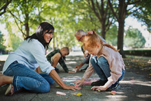 Senior Couple With Grandchildren Drawing With Chalks On Pavement Outdoors In Park.