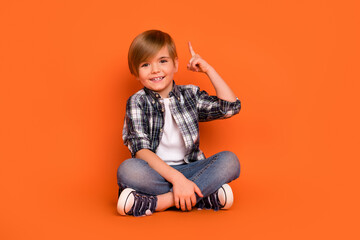 Full size photo of young boy sit floor indicate finger creative choice smart isolated over orange color background