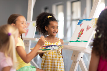 Wall Mural - Cheerful multiracial elementary schoolgirls standing by caucasian girl painting on easel in class