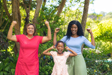 Wall Mural - Portrait of happy african american girl with mother and granddaughter flexing muscles in backyard