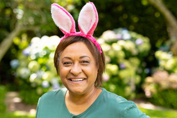 Wall Mural - Portrait of smiling african american senior woman wearing pink bunny ears in backyard