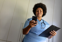 African American Mid Adult Female Nurse Using Smart Phone While Holding Clipboard In Hospital