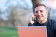 © Massimo Parisi - Portrait of attractive young office worker in casual wear, receives a phone call on his mobile phone, smiles and takes the call. I work remotely with the laptop from a table in the park.