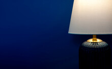 Close-up Luxury Table Lamp With White Fabric, Blue Glass, And Gold Color Materials Shining On Dark Blue Wall Background With Copy Space. Lighting Lamp In The Dark.