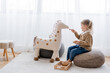 full length view of girl feeding toy horse from wooden bowl while playing on pouf at home.