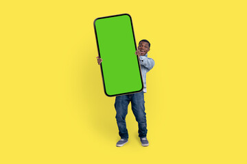 Excited black man standing and pointing Big smartphone with blank green screen, demonstrating copy space for app or website design, standing over yellow studio background, mockup image