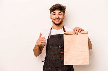 Wall Mural - Young hispanic clerk man holding a take away bag isolated on white background smiling and raising thumb up