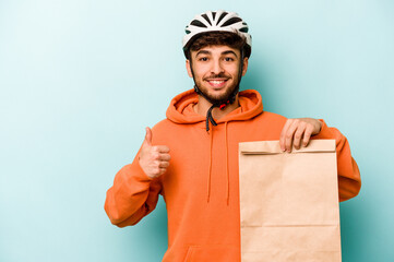 Wall Mural - Young hispanic man wearing a helmet bike holding a take away food isolated on blue background smiling and raising thumb up