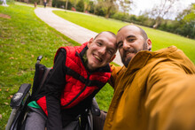 Selfie Of A Person With A Disability In A Wheelchair With A Friend On A Bench In A Public Park In The City, Talking And Laughing
