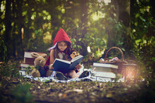 Books Can Transport Us To The Most Magical Places. Shot Of A Little Girl In A Red Cape Reading A Book With Her Toys In The Woods.