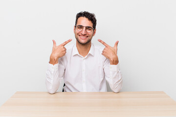 Wall Mural - hispanic handsome man smiling confidently pointing to own broad smile. businessman and desk concept