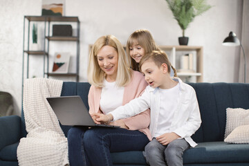 Wall Mural - Happy family of mother, little son and daughter sitting together on comfortable couch, using laptop, holding video call with father or greeting grandparents, recording video in living room.