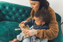 Lovely Curled Little Boy Sitting In His Mother's Lap Who Is Sitting On The Green Sofa And Looking At Something With His Mother Medium Full Shot Indoor Copy Space. High Quality Photo