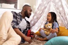 African American Man Talking To Cute Daughter Sitting With Teddy Bear In Tent At Home