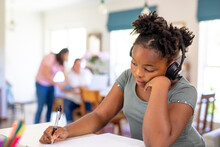 African American Girl With Headphone Writing On Book During Online School And Family In Background
