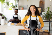 Portrait Of Smiling Young African American Female Barista Wearing Apron With Coworker In Background