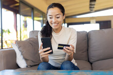 Happy Biracial Young Woman Holding Credit Card Using Smart Phone While Sitting On Sofa At Home