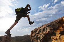 Low Angle View Of Athletic Young Male Caucasian Hiker Jumping On Rocky Cliffs During Sunny Day