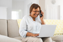 Happy African American Elderly Woman Talking On Smart Phone While Using Laptop At Home