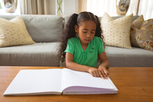 Biracial Girl Reading Book With Braille While Sitting In Living Room At Home