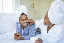 Happy Mother Applying Skin Cream On Nose Of Daughter In Bathrobe On Bed At Spa