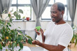 © pressmaster - Smiling African American pensioner in white t-shirt refreshening green leaves of domestic plant while spraying water on them