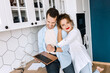 © shunevich - Happy family couple are standing at the kitchen at home, doing online shopping or chatting with friends. Joyful stylish husband and wife using laptop, watching funny videos or photos, smiling