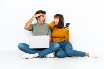 Wall Mural - Young couple with a laptop and mobile sitting on the floor with surprise and shocked facial expression