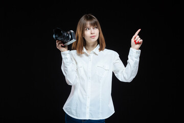 Portrait of a young woman wearing a white shirt, holding a camera in her hand and pointing with her hand at your text. The concept of a successful photographer. Isolated black background.