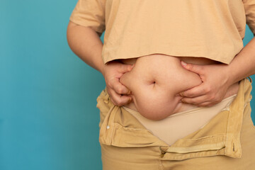 Cropped photo of plump plus-size overweight woman wearing beige T-shirt and jeans, showing squeezing excess naked belly on blue background. Body positive, cellulite, obesity, weight loss, liposuction.