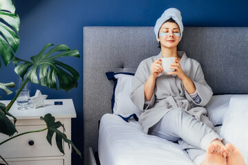 Woman in pajama and bathrobe with eyes patches and towel on her head relaxing and drinking morning coffee while sitting on her bed. Morning habits and home beauty routine. Self care. Selective focus