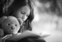 The Girl Is Reading A Story Book With A Teddy Bear.