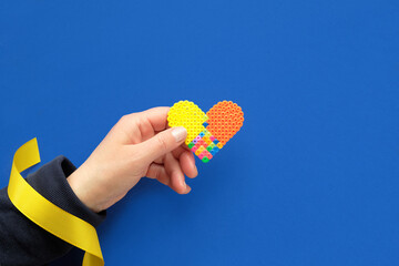 Wall Mural - Text Help Ukraine, hand with yellow ribbon holds multicolor plastic heart. Flat lay, top view on blue background. Volunteer help to refugees from Ukraine.