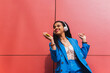 © Dani D.G - Exultant young woman Dancing and listening to music outdoors, wearing blue 90s clothes and red background. Happy girl of american ethnicity outdoors.