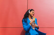 © Dani D.G - Woman singing with her mobile phone in an image of humor and fun while listening to music with her wireless headphones. Girl dancing outdoors on red background.