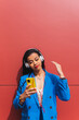 © Dani D.G - Young woman in fashionable blue clothes, relaxed, listening to music with her yellow smart phone on a red wall background. Image with commercial space.