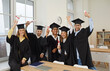 © Studio Romantic - Congrats, grad. Group of happy international multiracial multicultural male and female university graduate students in graduation hats and robes standing in classroom, cheering and raising up diplomas