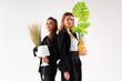 © Andrii - Two young business woman dressed black suit smile holding houseplant and lamp standing isolated on white background. Moving concept. Relocation new office with positive.