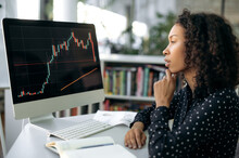Focused Smart African American Woman, Trader, Stock Broker, Sits At A Working Table In Front Of Computer Screen, Concentrated Looks At The Quotes Charts, Analyzes The Dynamics And Further Dynamics