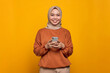 © Sewupari Studio - Smiling young Asian woman in orange shirt using mobile phone and looking at camera isolated over yellow background
