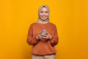 Wall Mural - Smiling young Asian woman in orange shirt using mobile phone and looking at camera isolated over yellow background