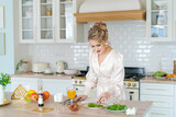 Fototapeta  - Beautiful woman cooking organic vegetarian salad, slicing strawberries and cheese in home kitchen interior. Girl with Vegan Food fresh ingredients, healthy cooking. In light dress with makeup and hair